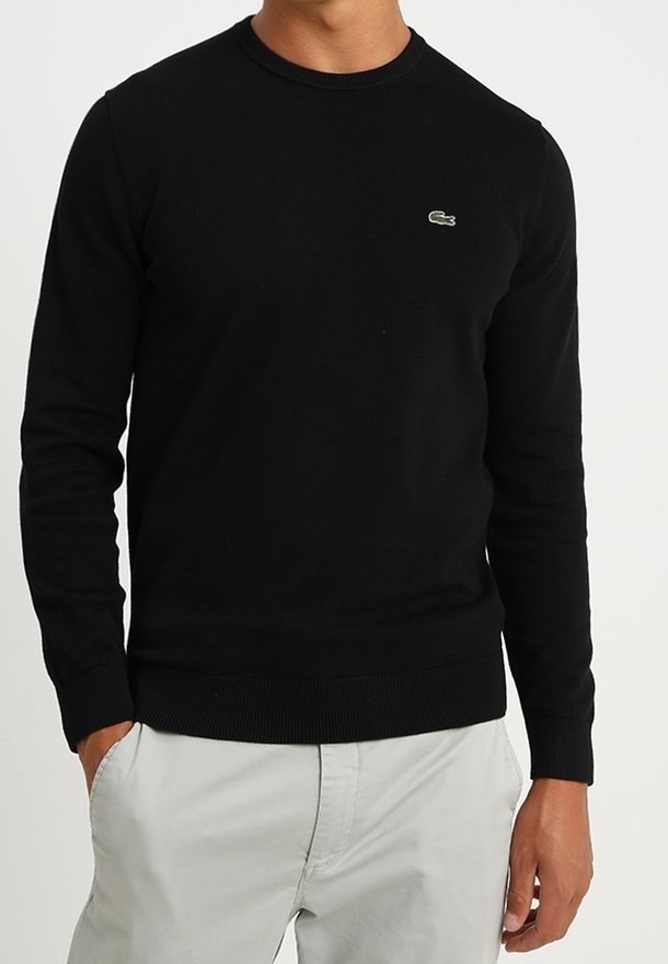 Un homme porte un pull noir à manches longues avec un petit logo de crocodile vert sur la poitrine, associé à un pantalon gris clair, les mains dans les poches.