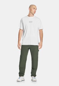 Biała bawełniana koszulka z małym szarym logo, zestawiona z oliwkowymi spodniami typu cargo z licznymi kieszeniami i luźnym krojem, noszona z białymi trampkami.