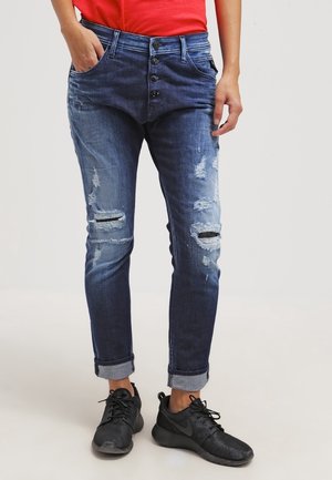 Osoba ubrana w podniszczone niebieskie dżinsy z podwiniętymi nogawkami, z odkrytym zapięciem na guziki, czerwoną bluzkę oraz czarne buty sportowe stoi.