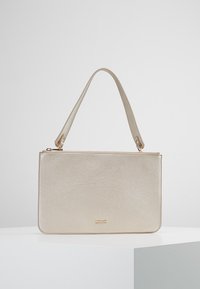 Petit sac à main rectangulaire en cuir beige avec une seule courte bandoulière et une fermeture éclair dorée sur le dessus, posé sur une surface blanche.