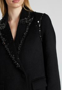 Abrigo de lana negro con solapas adornadas con lentejuelas oscuras, cierre de doble botonadura y bolsillos laterales. Diseño clásico y entallado.