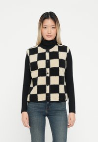 Gilet a righe nere e crema con una texture pelosa, collo rotondo e apertura con bottoni, indossato sopra una maglietta nera a maniche lunghe.