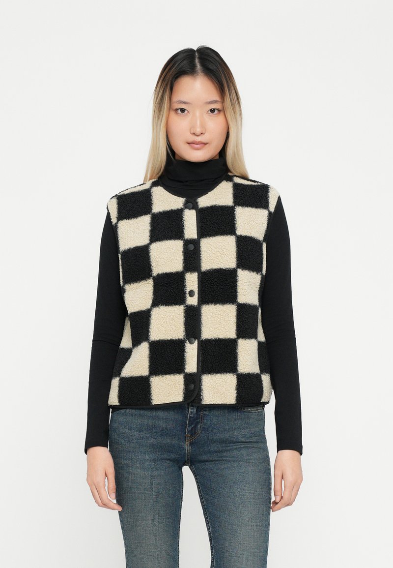 Gilet a righe nere e crema con una texture pelosa, collo rotondo e apertura con bottoni, indossato sopra una maglietta nera a maniche lunghe.