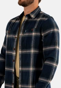 Camisa de franela en color marino con un patrón de cuadros en rojo y blanco, con cierres de botones y bolsillos en el pecho. Material: algodón suave.