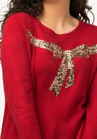 Czerwony sweter wykonany z miękkiej tkaniny posiada złotą kokardę z cekinów na przodzie, co podkreśla jego fakturę i atrakcyjność wizualną.