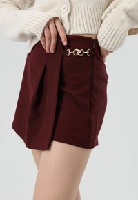 Bordeaux plissérok met een ritssluiting aan de zijkant en gouden kettingdetails, gemaakt van gladde stof. De plooien zorgen voor een gestructureerde vorm.