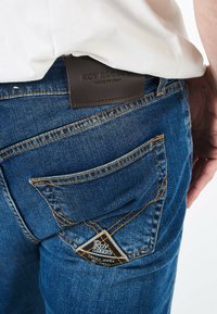 Jeans in denim blu con cuciture visibili, caratterizzati da una toppa triangolare in pelle nera sulla tasca posteriore e una texture liscia.