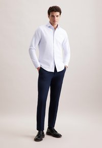 Camicia bianca con righe verticali, pantaloni slim-fit blu scuro e scarpe eleganti nere, modello in posa con le mani in tasca.