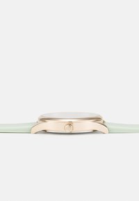 Gullfarget klokke med rund urskive, slank kasse og grønn silikonsnor. Har en enkelt krone på siden for justeringer. Minimalistisk design.