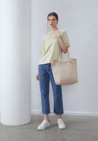 Geantă tote bej cu finisaj texturat, cu două bretele pentru umăr și noduri decorative; purtată cu un tricou galben deschis și blugi scurți albaștri.