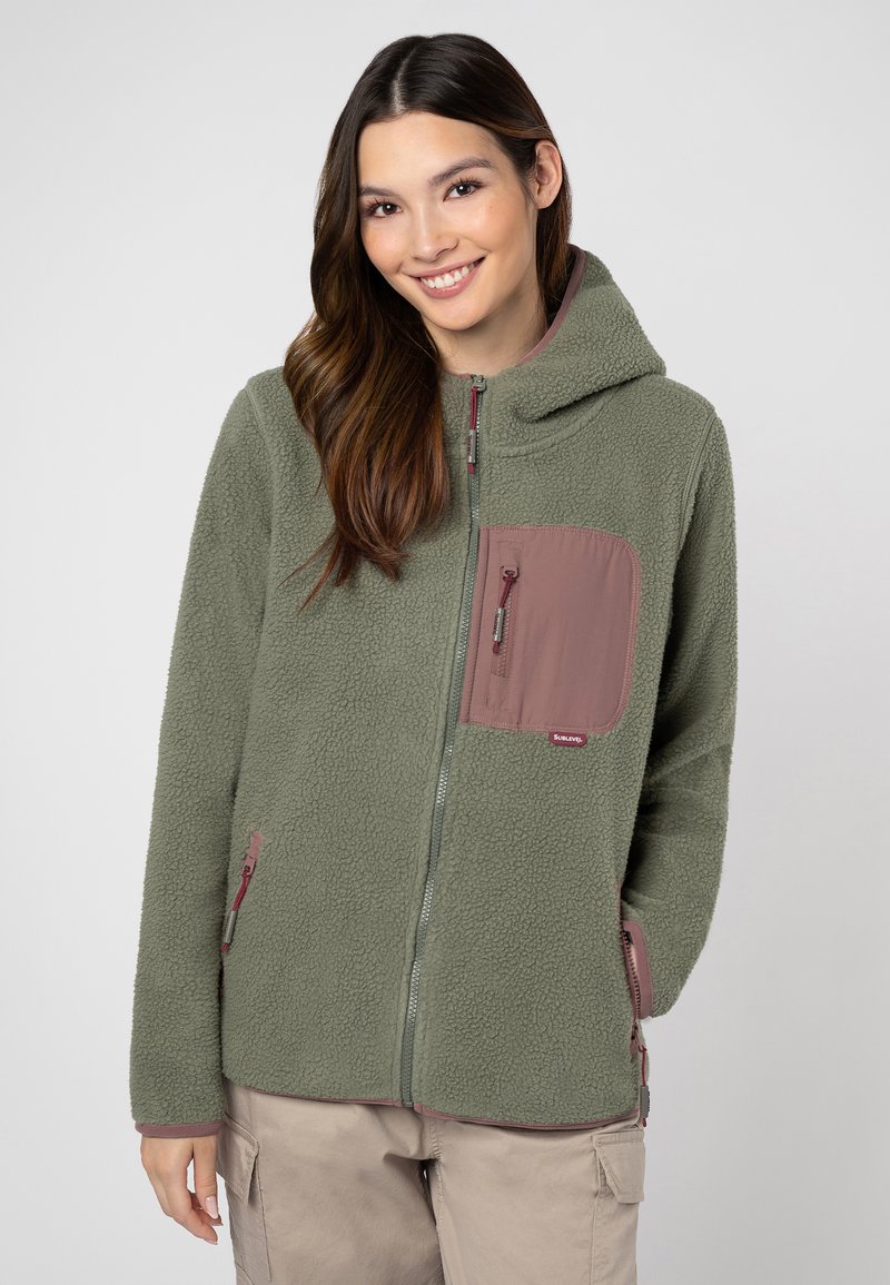 Jonge vrouw in een groene fleece jas met mauve ritszakken en beige cargobroek, glimlachend met haar handen in de zakken.