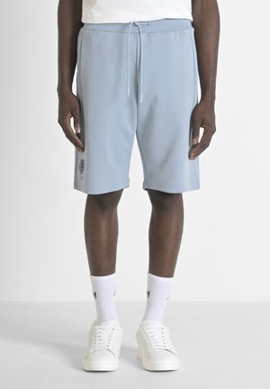 Shorts deportivos de color azul claro con cintura elástica y cordón de ajuste, que presentan un logo en el muslo izquierdo. Combinados con zapatillas blancas y calcetines.