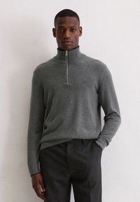 Grijze ritsvest van zacht stof met een hoge kraag, met ribbelboorden en -zoom. Draagt met een donkere, gestructureerde broek.