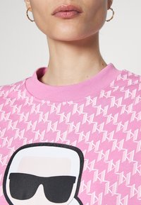 Różowa bluza z nadrukiem powtarzającego się wzoru logo oraz dużą grafiką postaci w okularach przeciwsłonecznych. Miękka tkanina i prążkowany dekolt.