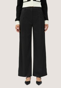 Zwarte wijde broek met een hoge taille, voorzien van scherpe plooien, een knoopsluiting en een gladde textuur. Gecombineerd met zwarte puntige schoenen.