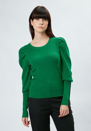 Suéter verde de punto con mangas abullonadas, escote en forma de barco y textura acanalada. Combinado con pantalones negros, que destacan una silueta ceñida.