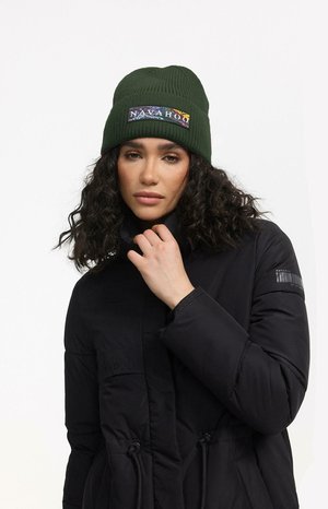 Mörkgrön ribbad mössa med en färgglad rektangulär lapp med texten "NAVAHOO." Modellen bär en svart puffjacka med hög krage.
