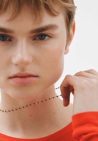 Collar dorado con cuentas negras y naranjas. La modelo lleva una camiseta de manga larga naranja, con piel clara y cabello castaño claro.
