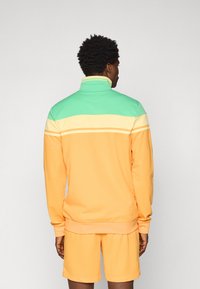 Lys orange genser med høy krage, grønne og gule striper over skuldrene, kombinert med matchende shorts. Myk tekstur.