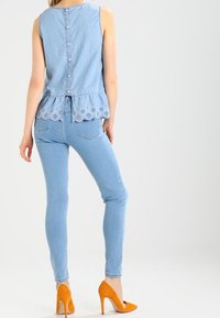 Haut sans manches en denim bleu clair avec un ourlet festonné et des détails en œillets, associé à un jean skinny bleu clair et des talons hauts orange.