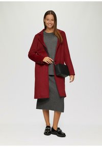 Czerwony wełniany płaszcz z wykładanym kołnierzem, szara dzianinowa sukienka i czarna torebka na crossbody. Model nosi czarne mokasyny; widoczne gładkie, teksturowane materiały.