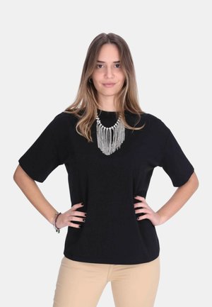 Donna con capelli lunghi in piedi con le mani sui fianchi, indossa una maglietta nera a maniche corte, pantaloni beige e una collana argentata con frange su uno sfondo semplice.