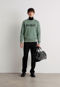 Grønn hettegenser med stor "HUGO" tekst, svarte smale bukser, svart lær helsvart bag, og svarte sko, plassert mot en ensfarget bakgrunn.