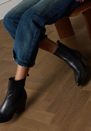 Schwarze Leder-Stiefeletten mit spitzer Zehenpartie und Blockabsatz, getragen zu hochgekrempelten blauen Jeans, auf einem strukturierten Holzboden.