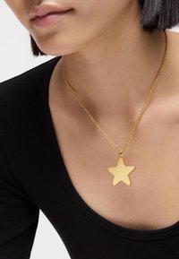 Collana a pendente a forma di stella dorata su una catena fine, indossata su un top nero, che mette in mostra una superficie liscia e un design minimalista.