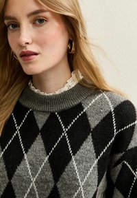 Joven mujer con cabello rubio que lleva pendientes de aro dorados y un suéter argyle gris y negro con un cuello de encaje blanco.
