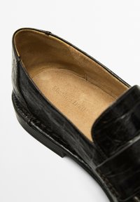 Black leather loafer with crocodile texture and tan suede insole showing brand embossing, viewed from above on a white background.