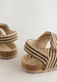 Flätade sandaler med beige och svart mönster, med en korksula och kardborreband för stängning. Mjuk tygfoder för komfort.