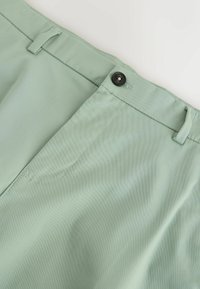 Pantalon vert clair avec une fermeture à bouton noir, des passants de ceinture et des coutures visibles sur les poches avant, en tissu lisse.