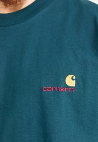 Teal katoenen t-shirt met een ronde hals. Bevat een geborduurd geel en roze Carhartt-logo op de linkerborst.