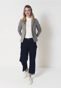 Šedý tvídový blazer přes bílý rolák, doplněný námořnickými širokými kalhotami a bílými teniskami, stojící na neutrálním pozadí.