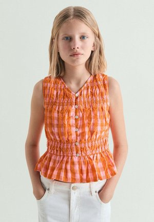 Niña con cabello rubio que lleva una blusa sin mangas a cuadros naranja y rosa con botones y pantalones blancos, de pie con las manos en los bolsillos.