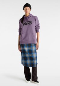 Felpa viola con logo "VANS", abbinata a una gonna maxi scozzese nei toni del blu e grigio. Il modello indossa scarpe nere e un berretto.