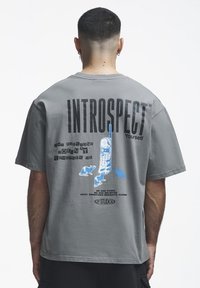 Homme aux cheveux courts portant un T-shirt gris imprimé "INTROSPECT Yourself", un téléphone et des souris, et un texte à propos d'être oublié et d'événements dramatiques.