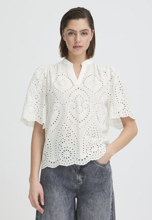 Femeie purtând o bluză scurtă cu mâneci din dantelă albă tip ochi de plasă, cu margini ondulate și blugi gri închis cu talie înaltă, cu mâna în buzunar.