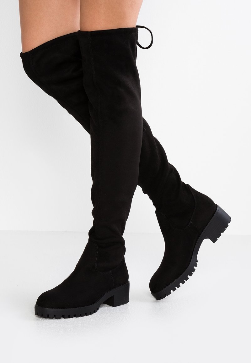 Bottes noires en daim au-dessus du genou avec semelles crantées épaisses portées sur des jambes croisées sur un fond blanc uni.