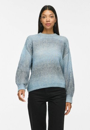 Kvinde med langt mørkt hår iført en blå gradientstrikket sweater og sorte bukser, stående foran en ensfarvet hvid baggrund.
