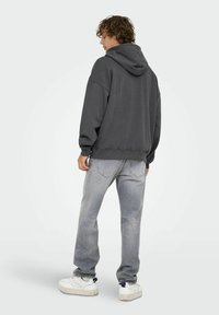Joven con cabello rizado que lleva una sudadera gris oscuro de gran tamaño, jeans gris claro y zapatillas blancas, de pie con un ligero giro hacia un lado.