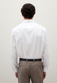 Camicia bianca a maniche lunghe, colletto classico e tessuto texturizzato, abbinata a pantaloni marrone chiaro e una cintura nera.
