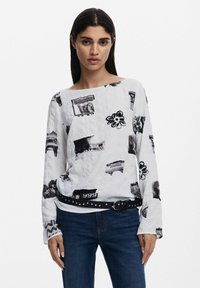 Top alb cu maneci lungi, având imprimeuri grafice negre, inclusiv flori și diverse forme, realizat dintr-un material texturat și cu o curea neagră cu ținte.