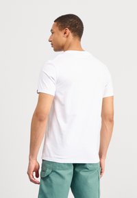 Weißes T-Shirt mit kurzen Ärmeln, runder Halslinie, glatter Textur und dezentem Branding am Ärmel; getragen mit grünen Hosen.