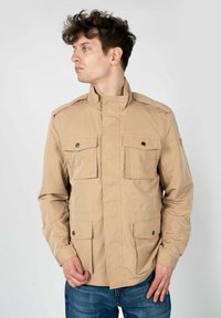 Giacca utility beige con colletto alto, due tasche sul petto e due tasche laterali. Presenta un tessuto leggero e testurizzato con dettagli a bottoni.