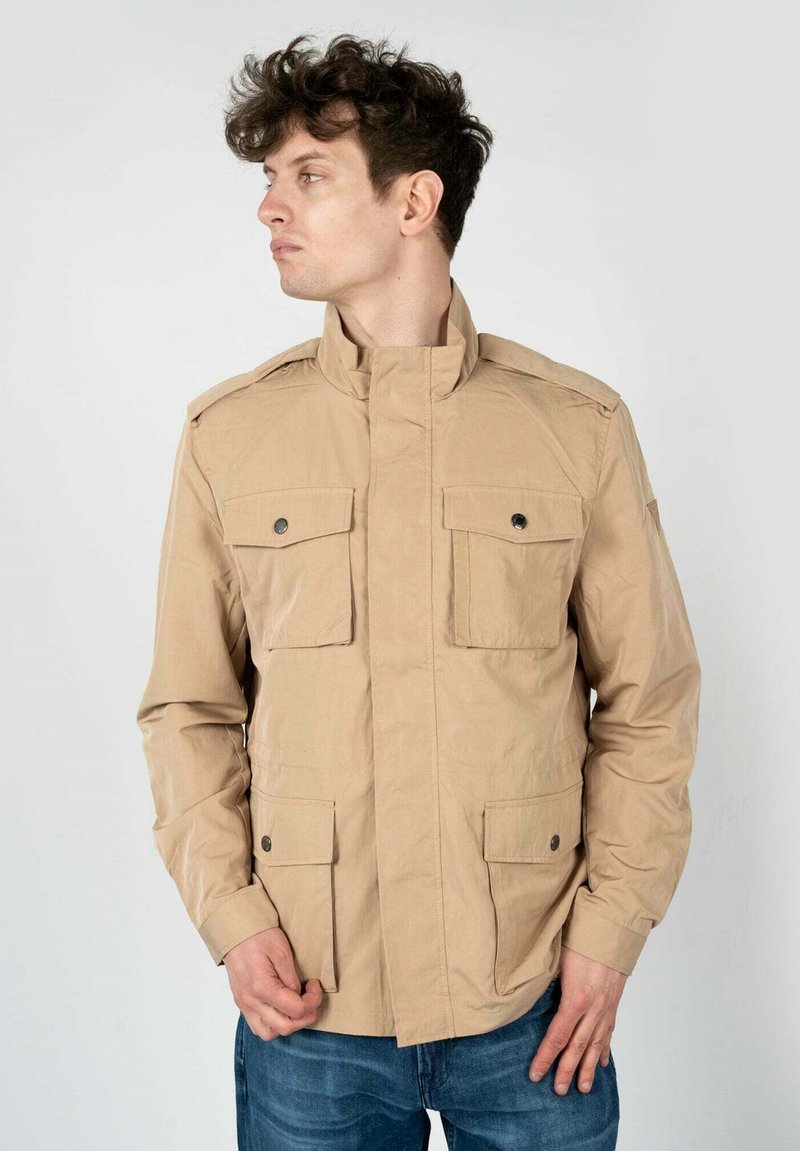 Giacca utility beige con colletto alto, due tasche sul petto e due tasche laterali. Presenta un tessuto leggero e testurizzato con dettagli a bottoni.