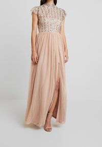 Lång, rosafärgad klänning med en paljettprydd överdel, kortärmade ärmar och en hög slits på låret. Tyget är genomskinligt och flödande, vilket framhäver silhuetten.