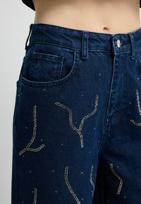 Vaqueros de mezclilla azul oscuro con diseño de cintura alta, bolsillos y patrones intrincados de tachuelas plateadas en toda la tela.