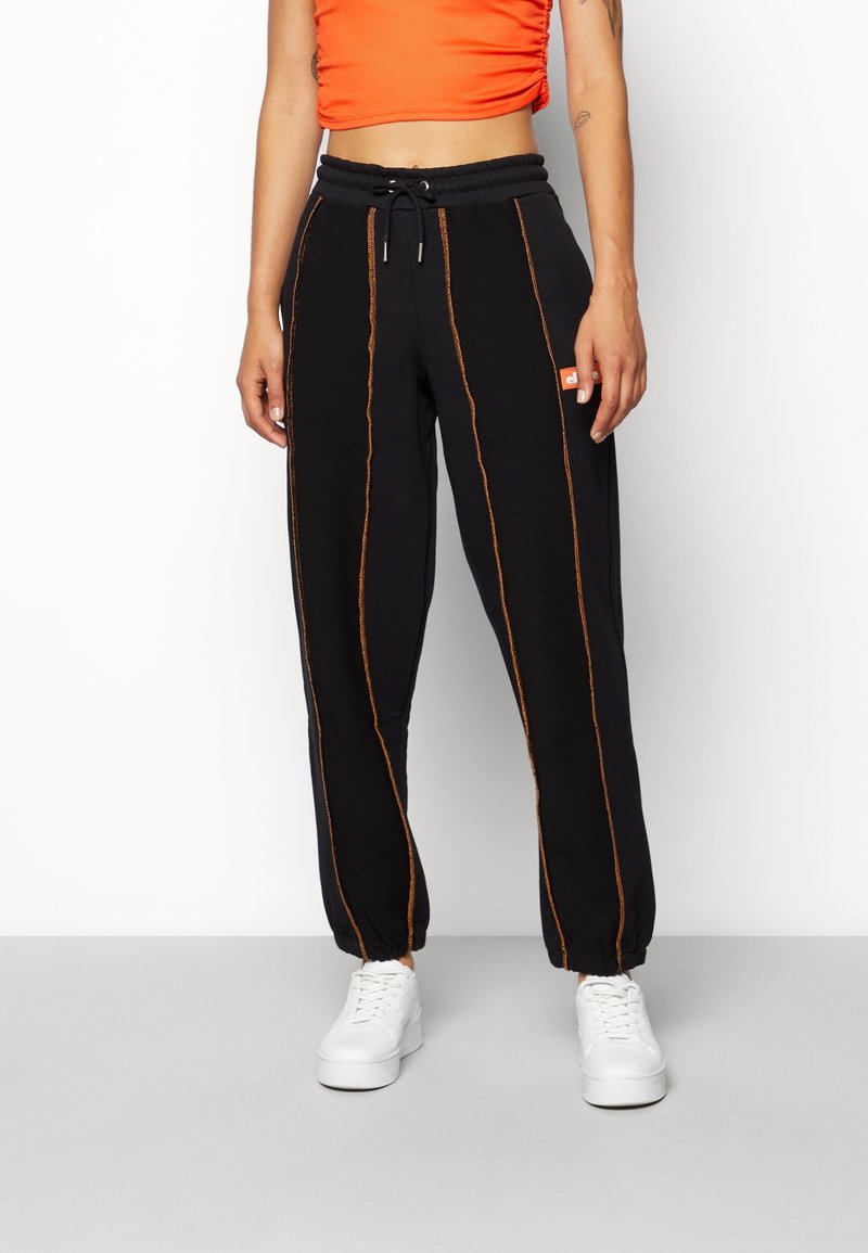 Close-up van een persoon die zwarte joggingsbroeken met oranje verticale naden, een bijpassend oranje cropped top en witte sneakers draagt, op een effen achtergrond.